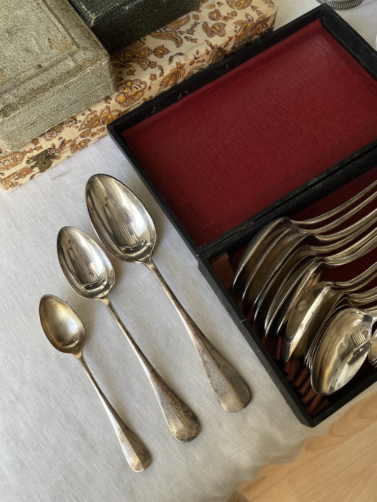 A vintage set of silver spoons and forks arranged on a linen tablecloth beside old books.